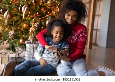 Happy African American woman with adorable little daughter using phone, looking on smartphone screen, shopping online, choosing gifts, sitting on warm floor near Christmas tree at home - Powered by Shutterstock