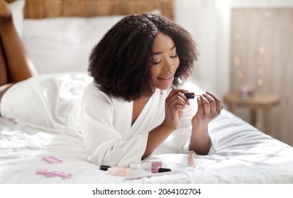 Happy African American Woman Applying Nail Polish, Making Manicure, Lying On Bed In Dressing Gown, Doing Domestic Spa Procedure. Beautiful Black Lady Taking Care Of Herself At Home