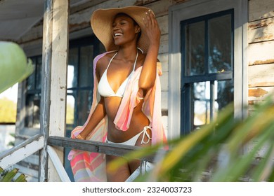 Happy african american woman in bikini and sunhat standing on porch of beach house, copy space. Summer, fashion and beauty, free time and vacations, unaltered. - Powered by Shutterstock