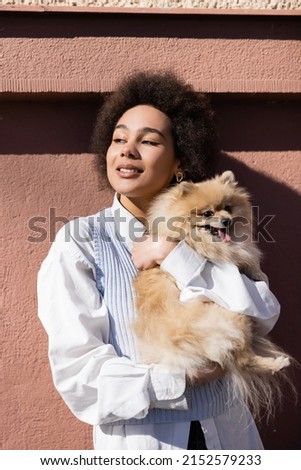 happy african american woman in blue vest holding in arms pomeranian spitz near wall