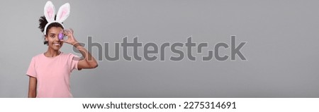 Happy African American woman with bunny ears holding Easter egg near eye on light grey background, space for text. Banner design
