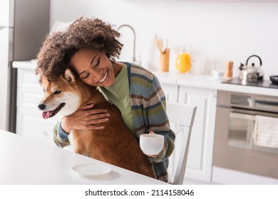 happy african american woman with closed eyes and coffee cup embracing shiba inu dog in kitchen - Powered by Shutterstock