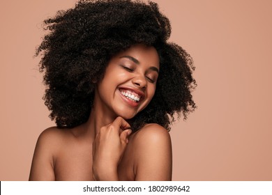 Happy African American woman with curly hair closing eyes and cheerfully smiling while enjoying clean skin after spa session against brown background