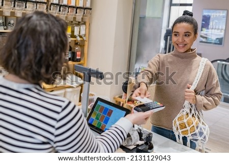 Happy african american woman with eco string bag making payment with credit card on POS terminal in sustainable local shop standing before counter. Woman at cashdesk paying purchase at grocery shop