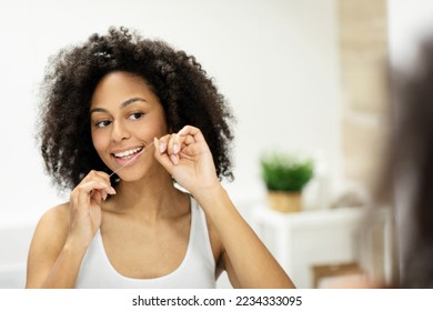 Happy african american woman flossing her teeth with dental floss in front of mirror at home, enjoying her perfect white smile