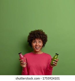 Happy African American Woman Has Healthy Meal, Holds Strawberry And Spinach Smoothie In Glass Bottles, Drinks Detox Beverage, Keeps To Diet, Leads Healthy Lifestyle, Concentrated Above On Empty Space