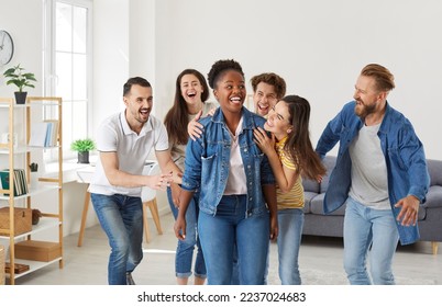 Happy African American woman having fun with several friends at a party at home. Group of cheerful joyful young people hug their friend as they laugh at her joke or congratulate her on great success - Powered by Shutterstock