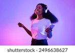 Happy African American woman with headphones and smartphone listening to music and dancing in neon light. Young black woman moving to favorite song, enjoying cool playlist