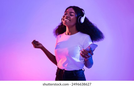 Happy African American woman with headphones and smartphone listening to music and dancing in neon light. Young black woman moving to favorite song, enjoying cool playlist - Powered by Shutterstock