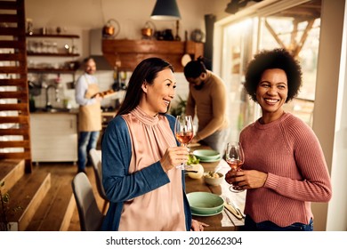 Happy African American Woman And Her Asian Female Friend Drinking Wine While Their Husbands Are Preparing Dinner In The Background. 