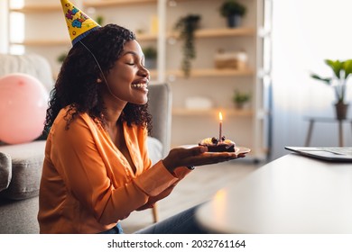 Happy African American Woman Holding Birthday Cake With Burning Candle And Making Wish Celebrating B-day Alone, Wearing Festive Hat Sitting At Home. Side View. Remote Holiday Celebration