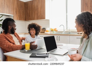 Happy African American Woman Holding Cup Near Laptop While Husband And Daughter Talking Near Digital Tablet In Kitchen