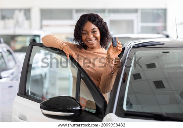 Happy African American Woman Holding Keys To New\
Car And Smiling At Camera, Excited Black Female Posing Near Her\
Vehicle In Dealership Center Or Rental Office, Leaning On\
Automobile Door, Free\
Space