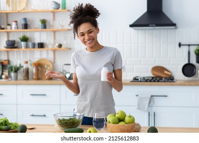 Happy african american woman in home kitchen holding a bottle of nutritional supplements, looking at the camera and smiling friendly, healthy lifestyle - Shutterstock ID 2125612883