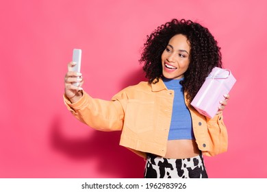 happy african american woman in jacket holding present while taking selfie on smartphone on pink - Powered by Shutterstock