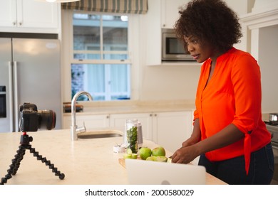 Happy African American Woman In Kitchen Preparing Health Drink, Making Vlog Using Laptop And Camera. Online Cookery Vlogger At Home.