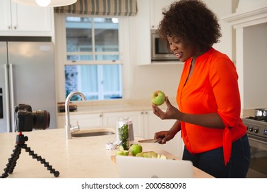 Happy African American Woman In Kitchen Preparing Health Drink, Making Vlog Using Laptop And Camera. Online Cookery Vlogger At Home.