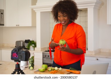 Happy African American Woman In Kitchen Preparing Health Drink, Making Vlog Using Laptop And Camera. Online Cookery Vlogger At Home.