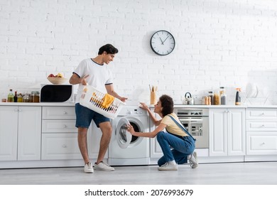 happy african american woman looking at boyfriend holding basket with dirty laundry near washing machine in kitchen - Powered by Shutterstock