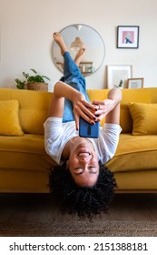 Happy African American woman lying upside down on the couch texting using mobile phone. Checking social media. Vertical.