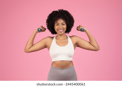 A happy African American woman with natural hair holds dumbbells up, showcasing fitness on a pink background - Powered by Shutterstock