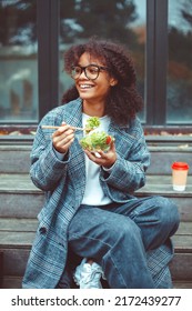 Happy African American Woman Office Worker Eating Salad And Smiling At Camera While Sitting On Bench In Park Outdoors, Selective Focus. Positive Black Girl Having Lunch Outside During Break At Work