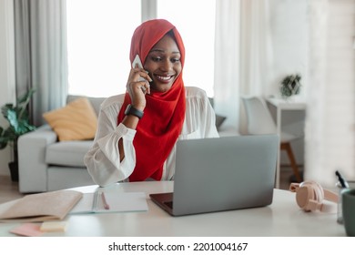 Happy african american woman in red hijab working on laptop at home office, looking at computer screen and talking on smartphone with client, copy space - Powered by Shutterstock