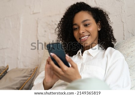 Happy african american woman relaxing on comfortable couch, holding smartphone in hands. Smiling young lady chatting in social networks, watching funny videos, using mobile applications at home.