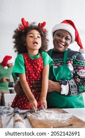 Happy African American Woman In Santa Hat Embracing Granddaughter While Cooking In Kitchen Together