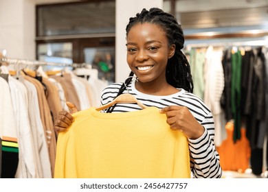 Happy African American Woman Shopping In Boutique, Holding A Bright Yellow Sweater, Smiling Confidently At The Camera In A Fashion Store - Powered by Shutterstock