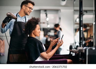 Happy African American Woman Showing Something On Smart Phone To Her Hairdresser While Getting Her Hair Styled At The Salon. 