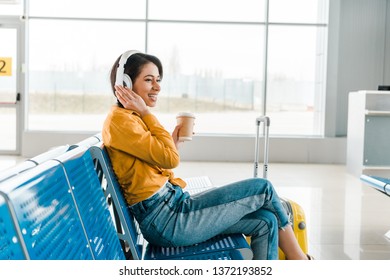 Happy African American Woman Sitting In Departure Lounge With Coffee To Go And Suitcase While Listening Music In Headphones 