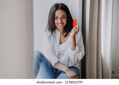 Happy African American Woman Sitting On Windowsill Looking At Camera Holding Credit Card, Wearing White Shirt And Blue Jeans At Home. Satisfied Afro Brazilian Girl Earned Money To Buy A New Home, Car.