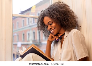 Happy african american woman sitting on the windowsill and reading a book.