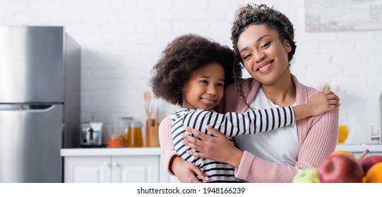 happy african american woman smiling at camera while embracing child in kitchen, banner