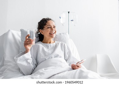 happy african american woman smiling while holding glass of water and mobile phone in clinic