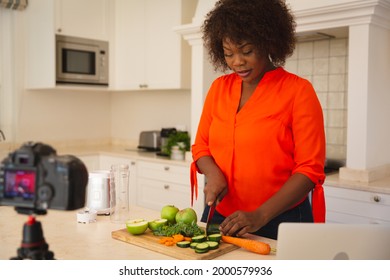 Happy African American Woman Standing In Kitchen Preparing Food, Making Vlog Using Laptop And Camera. Online Cookery Vlogger At Home.