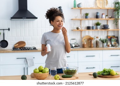 Happy african american woman standing at the cuisine table in the home kitchen drinking dietary supplements, looking away and smiling friendly, healthy lifestyle concept - Powered by Shutterstock