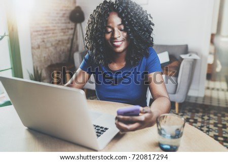 Happy african american woman using laptop and smartphone while sitting at wooden table in the living room.Horizontal.Blurred background