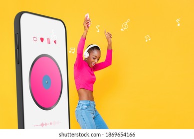Happy African American woman wearing headphones listening to streaming music from smartphone application and dancing on colorful yellow isolated studio background