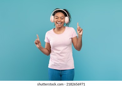 Happy African American Woman In Wireless Headphones Listening To Popular Music And Dancing On Blue Studio Background. Joyful Black Female Enjoying Favorite Song