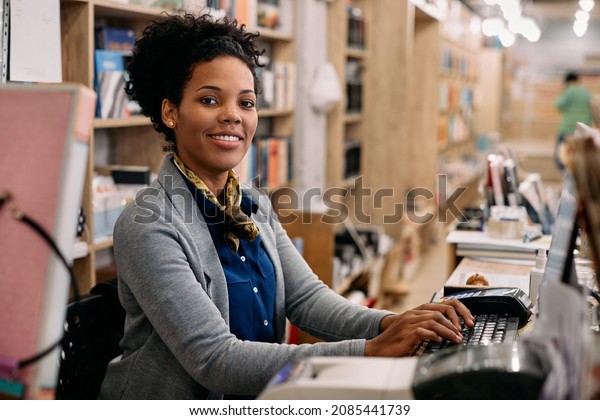 Happy African American woman working\
on a computer at bookstore and looking at camera.\
