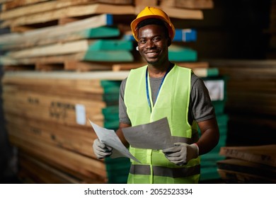 Happy African American Worker Going Through Delivery Plans At Lumber Distribution Warehouse And Looking At Camera.