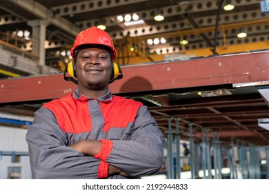 Happy African American Worker In Personal Protective Equipment Looking At Camera. Portrait Of Black Industrial Worker In Red Helmet, Hearing Protection Equipment And Work Uniform In A Factory.