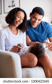 Happy African American Young Adult Parents Playing With Their Three Month Old Baby Son, Lying On His Mother’s Knee, Vertical
