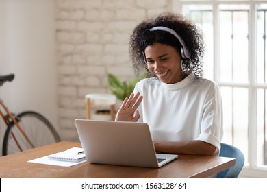 Happy african american young business woman wearing wireless headphones, looking at computer screen, waving hello. Pleasant attractive mixed race female professional holding video call with client.