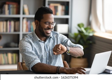 Happy African American Young Businessman Employee Worker Looking At Smart Watch Sit At Office Desk With Laptop, Smartwatch Digital Modern Technology Apps For Planning Work Time Management Concept