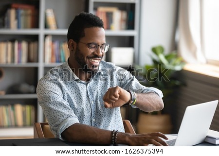 Happy african american young businessman employee worker looking at smart watch sit at office desk with laptop, smartwatch digital modern technology apps for planning work time management concept