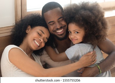 Happy african American young couple with preschooler sit hug posing looking at camera, loving biracial millennial parents with cute little daughter cuddle embrace enjoying tender moment at together