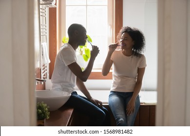Happy african American young couple sit in bathroom clean teeth with toothbrushes enjoy morning together, smiling loving biracial husband and wife perform daily cleanup routine in home bath